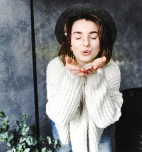 brunette woman in black hate with white sweater blowing snow at camera