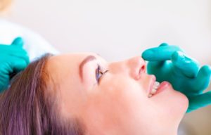 side profile of brunette woman's face laying down with doctors hands wearing blue gloves touching her face