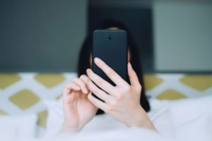 brunette woman laying in bed with a yellow and white headboard using black cell phone that blocks her face