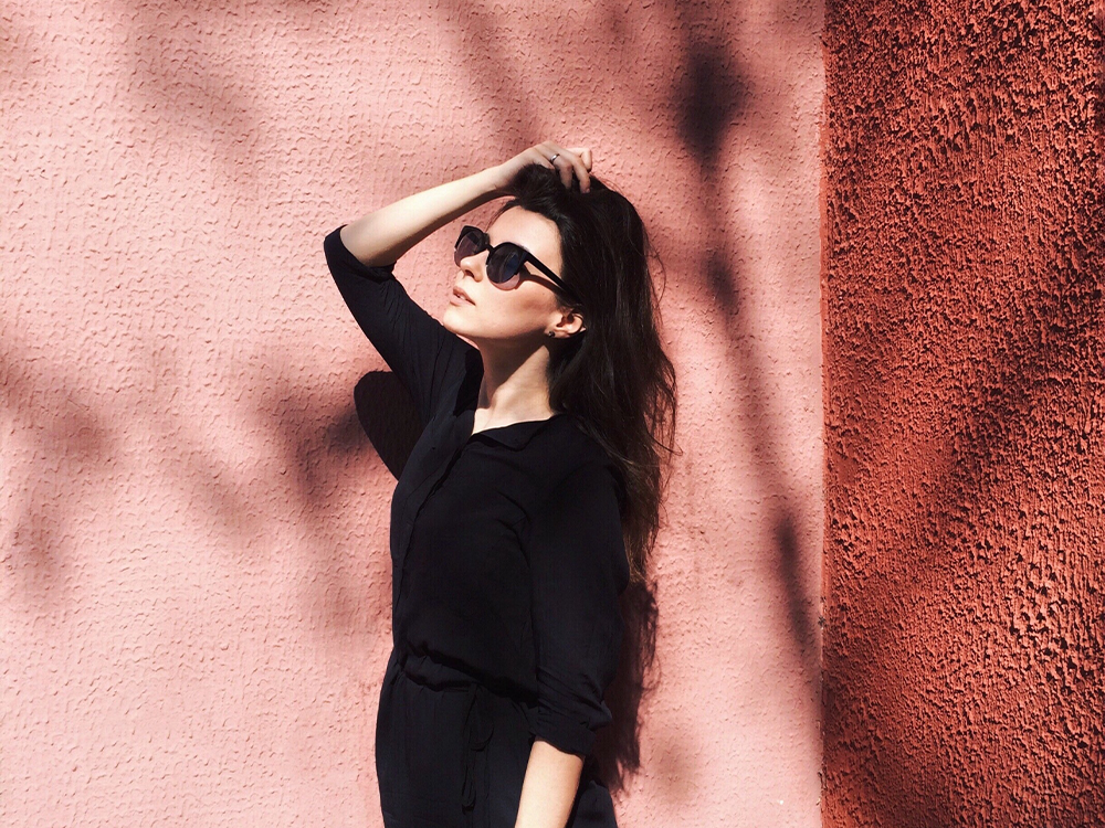 Brunette woman posing in front of pink wall, wearing black.