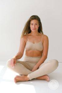 Woman sitting cross legged on a white background wearing a tan workout set