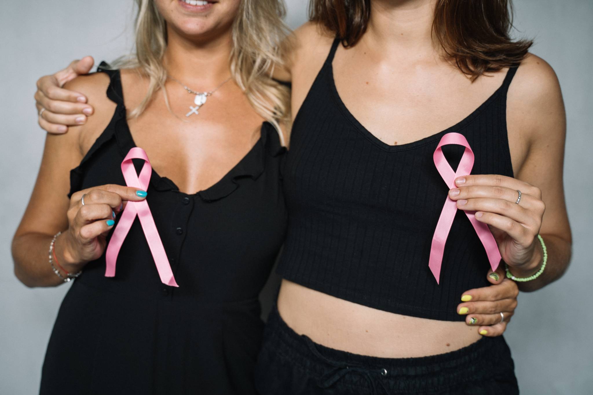 Blonde and brunette woman standing side-by-side wearing black T-shirts holding pink ribbons for breast cancer awareness