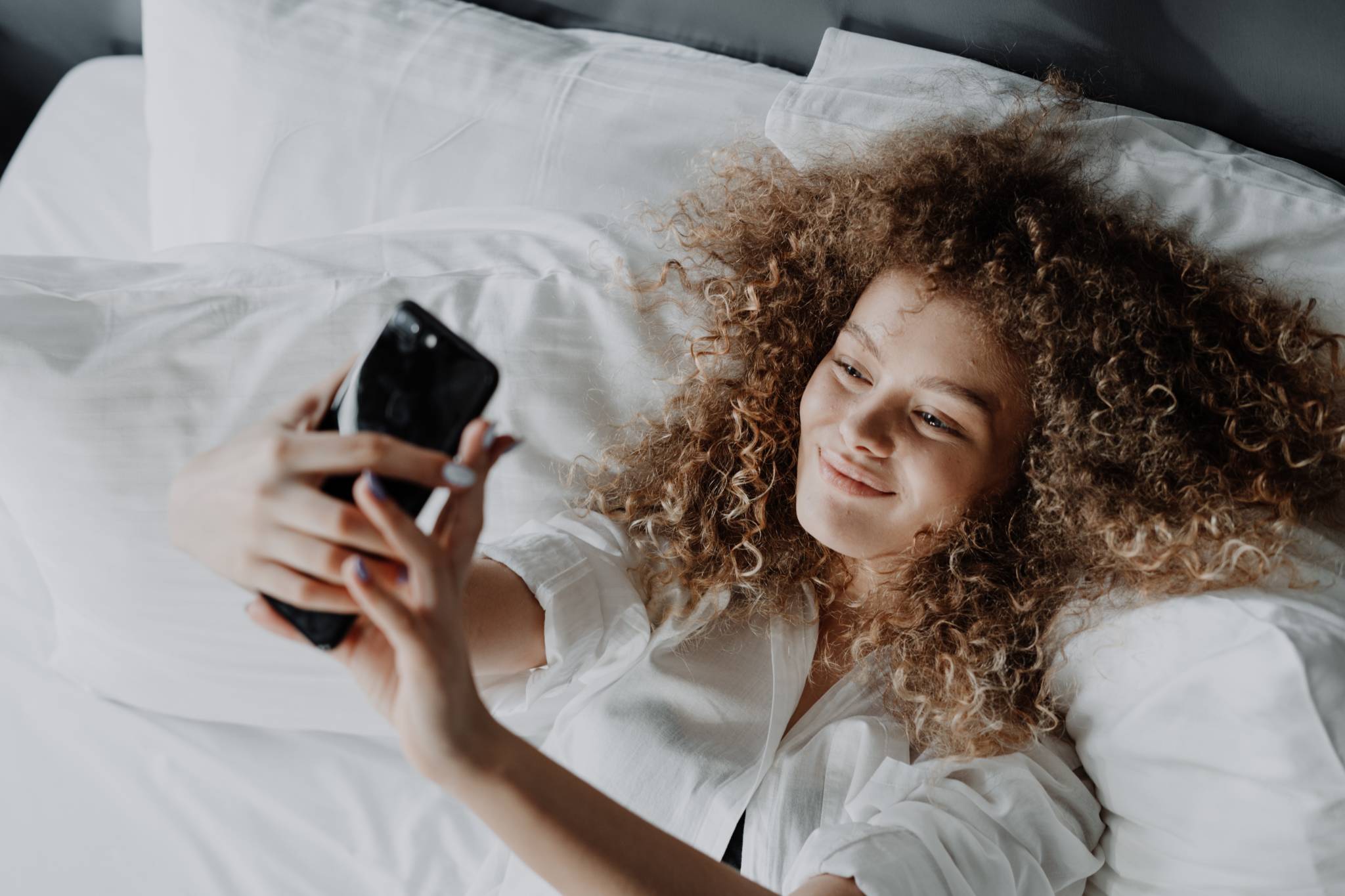 woman with curly hair lying in bed with white sheets and pillow cases taking a selfie smiling at smart phone above her head with outstretched arms