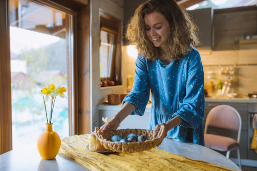 woman with curly hair wearing blue shirt placing basket of blue easter eggs on kitchen table
