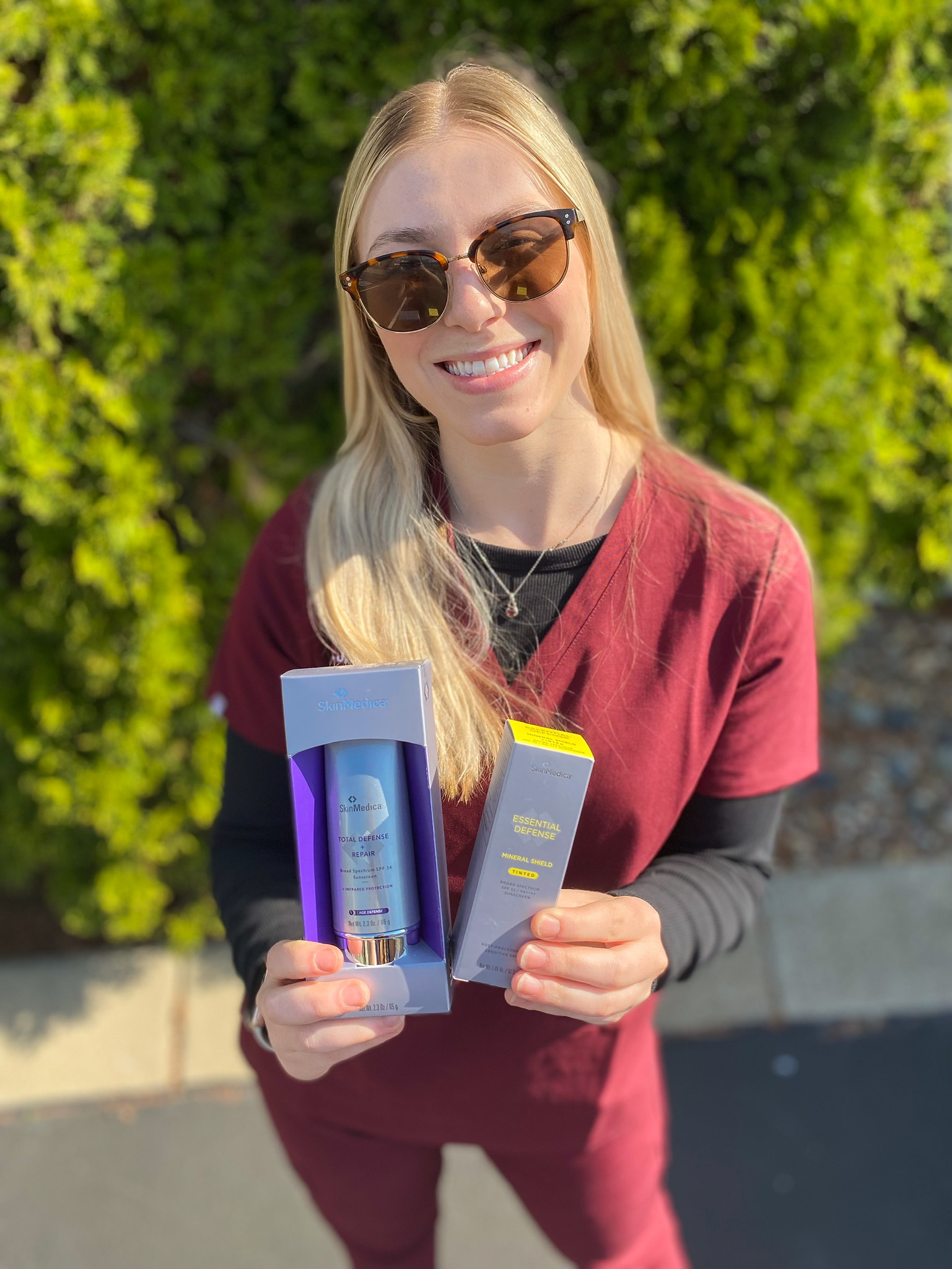 dr j j wendel employee holding up skinmedica sunscreen products while wearing scrubs outside in front of a hedge