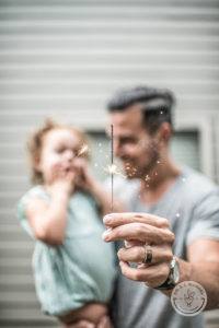 man holding young girl in right arm holding up a lit sparkler in his left hand