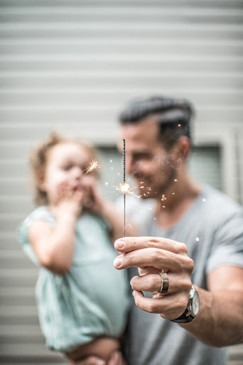 man holding young girl in right arm holding up a lit sparkler in his left hand