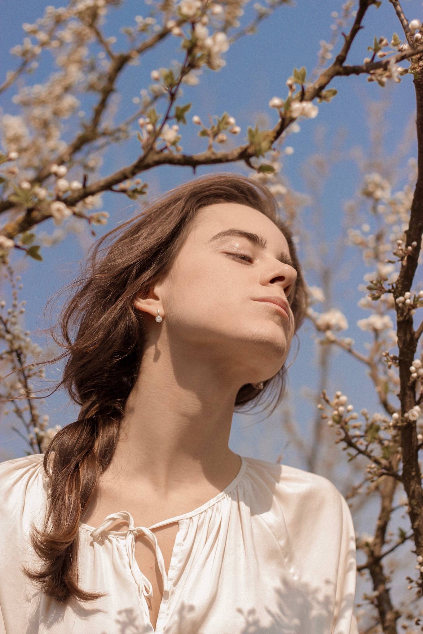 woman with braided hair closing eyes with chin turned up to sky, blue sky and flowering tree in background