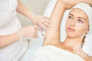 woman laying on white bed wearing white towel and headband while nurse injects botox for hyperhidrosis into armpit while wearing gloves