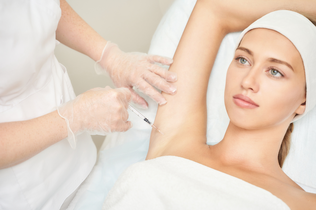 woman laying on white bed wearing white towel and headband while nurse injects botox for hyperhidrosis into armpit while wearing gloves