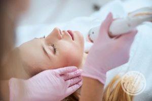 woman in white laying on table receiving microneedling treatment by doctor wearing pink surgical gloves