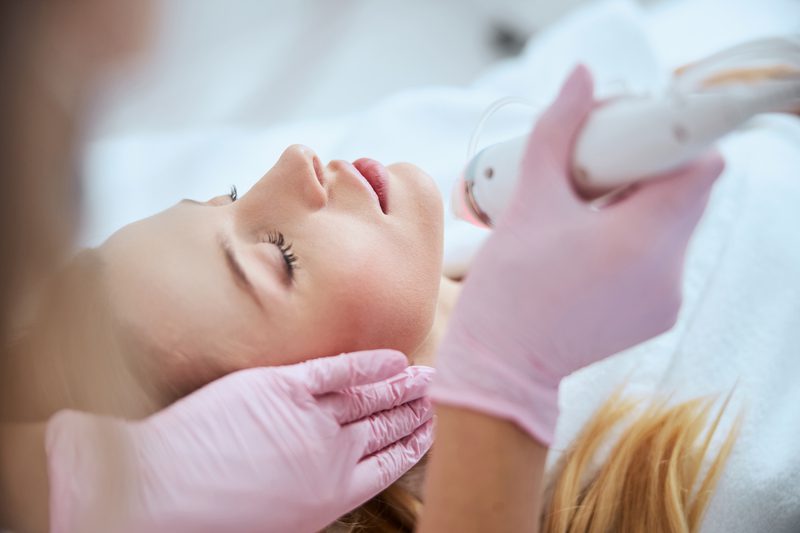 woman in white lying on table receiving microneedling treatment by doctor wearing pink surgical gloves