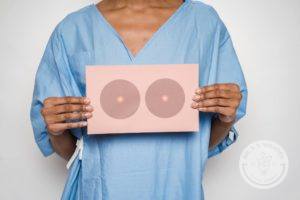 woman wearing light blue medical gown and holding pink card with a drawing of breasts on the front