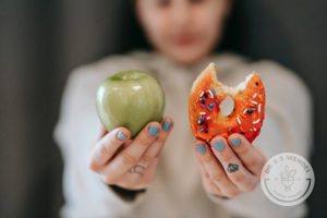 woman with light blue nails holds green apple and donut with orange icing and sprinkles