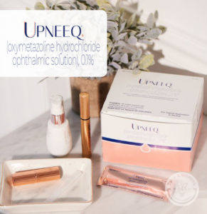 box of Upneeq eye drops and a sample eye drops next to lipstick and cosmetics on a table