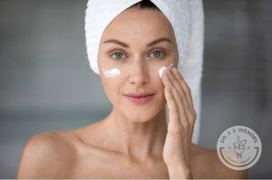 woman with hair wrapped in towel applies skincare to face