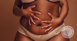 black woman in undergarments holds abdomen with both hands in front of brown background