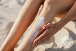 Woman sitting on white sand beach applying sunscreen to leg