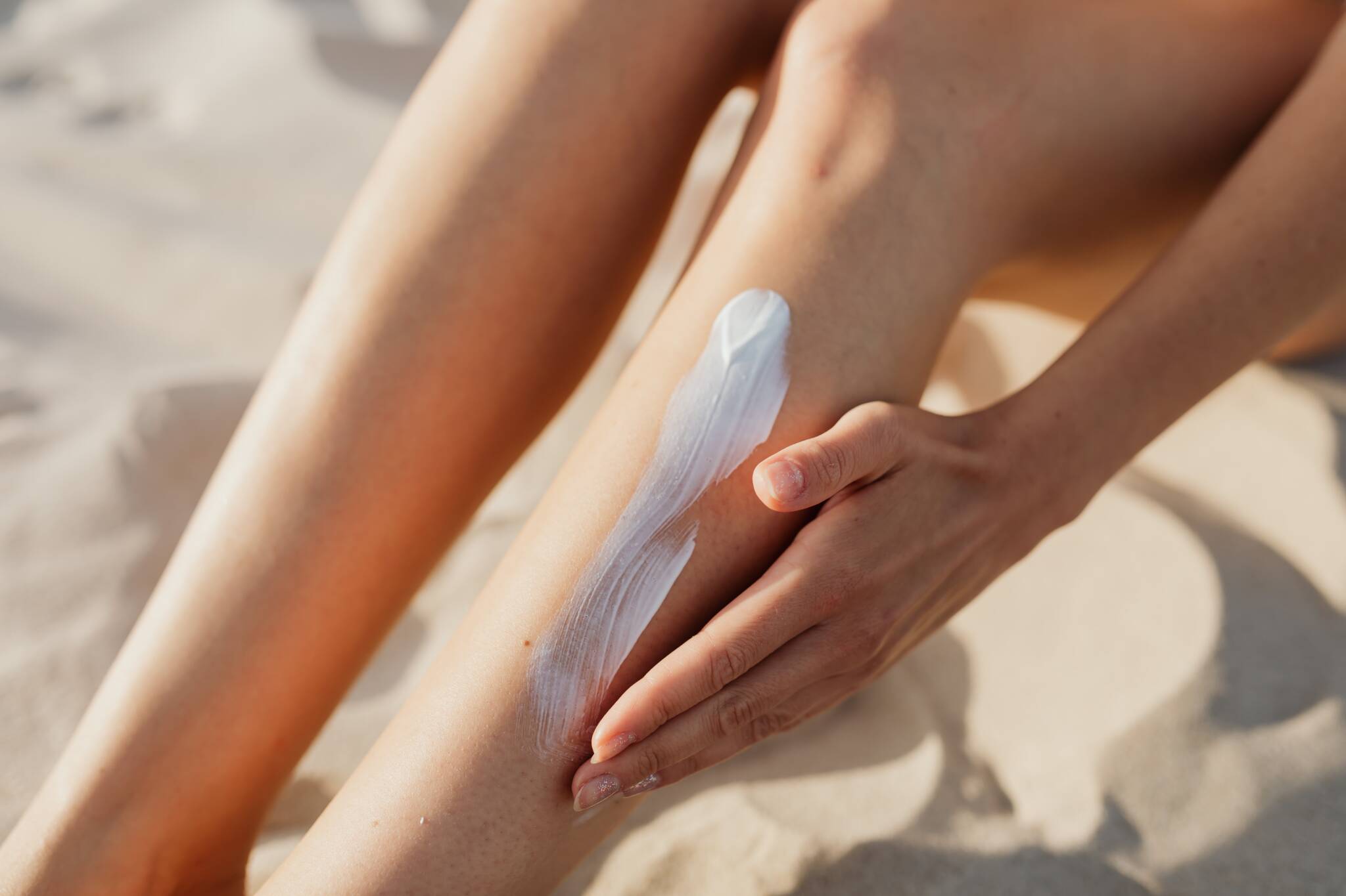 Woman sitting on white sand beach applying sunscreen to leg