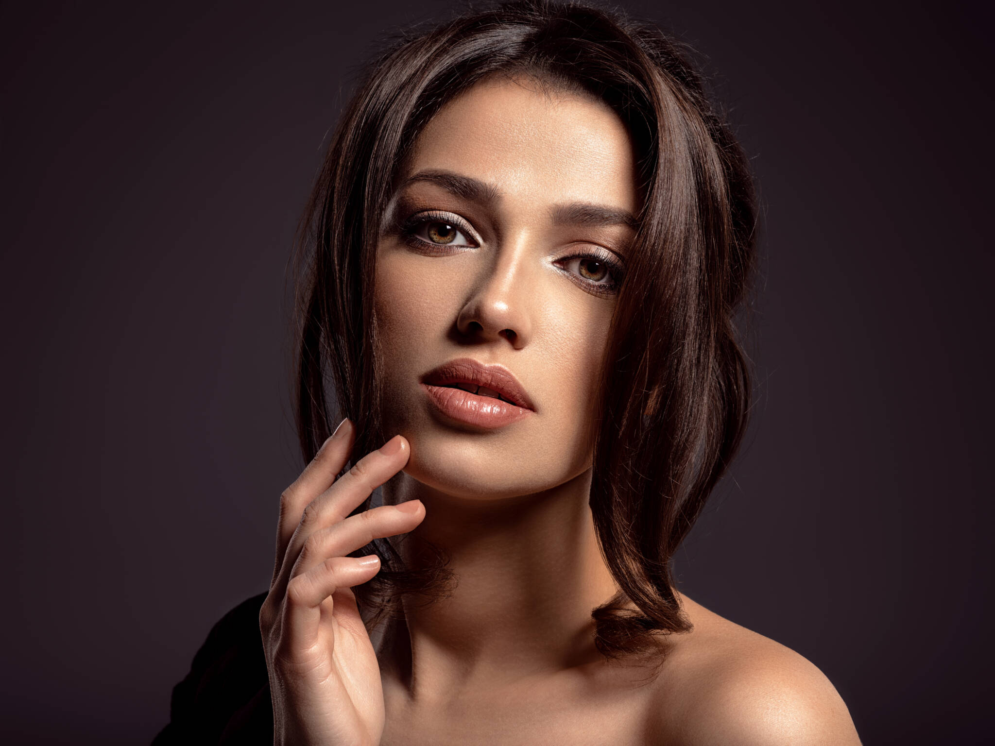 Woman with dark brown hair touching her chin in front of a brown backdrop