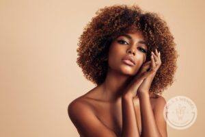 woman with curly hair against a tan background with hands placed on cheek