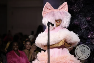 Sia wearing pink wig and giant bow in a frilly dress while standing in front of a mic