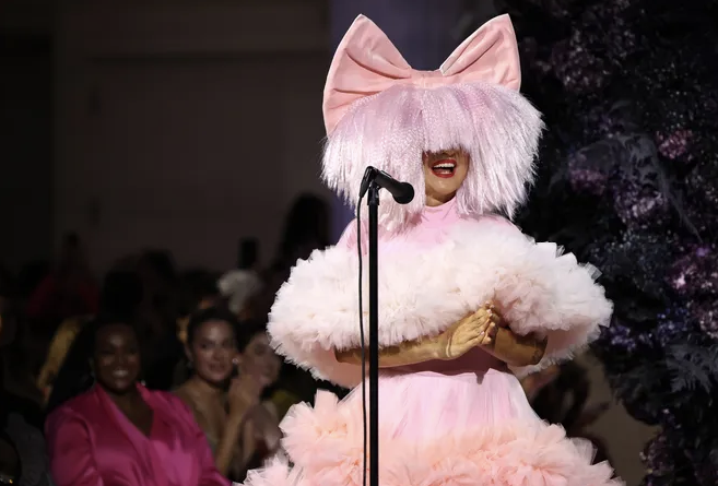 Sia wearing pink wig and giant bow in a frilly dress while standing in front of a mic