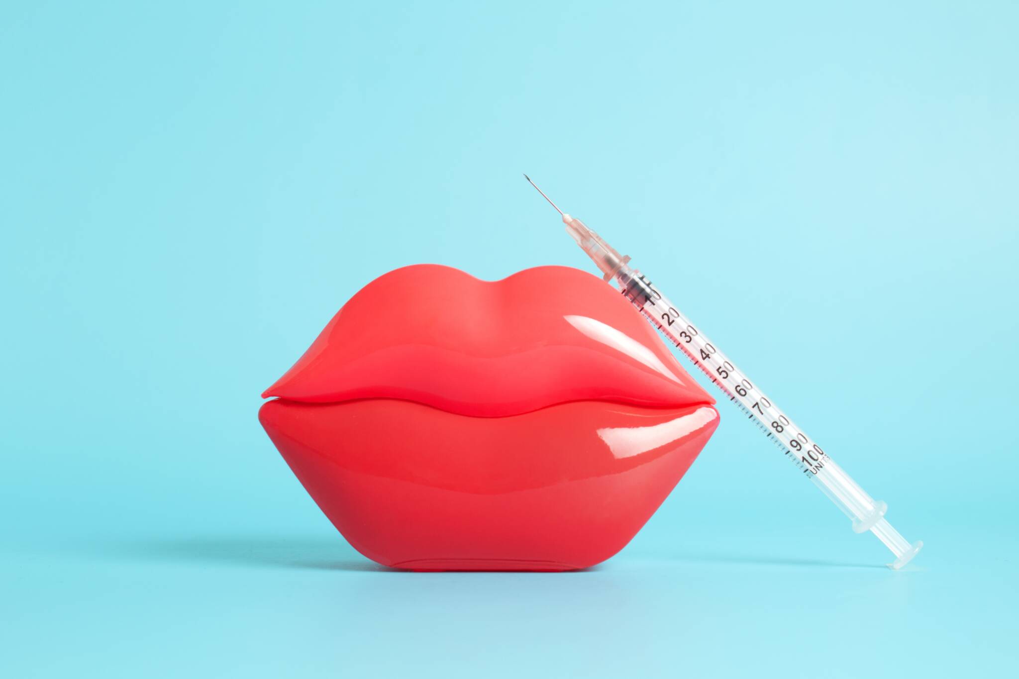 a syringe leaning against bright red plastic lips in front of a blue background