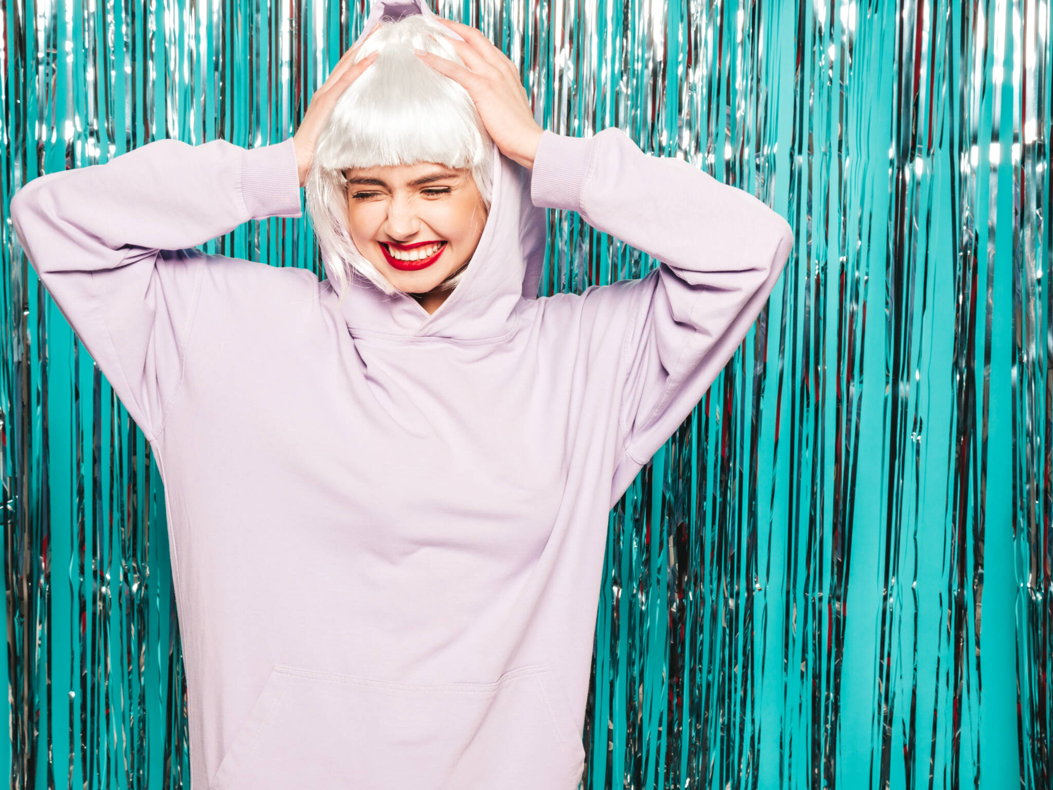 Young woman smiling while wearing a pink sweatshirt and platinum wig in front of teal background