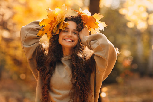 Brunette woman in brown sweater smiling while holding orange leaves against her head