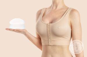 Woman holds round implants wearing compressing bra on beige background.