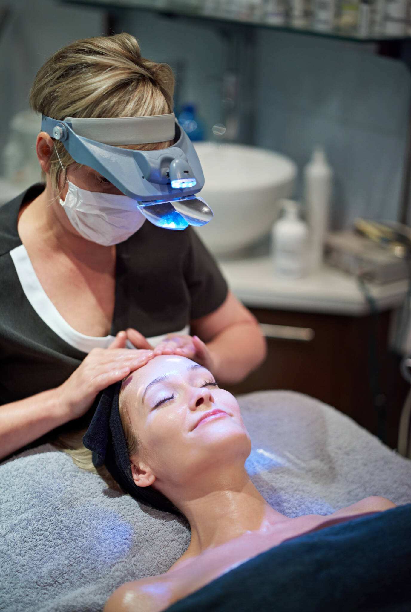 Woman receiving facial treatment by an esthetician wearing a mask and magnifying head lamp
