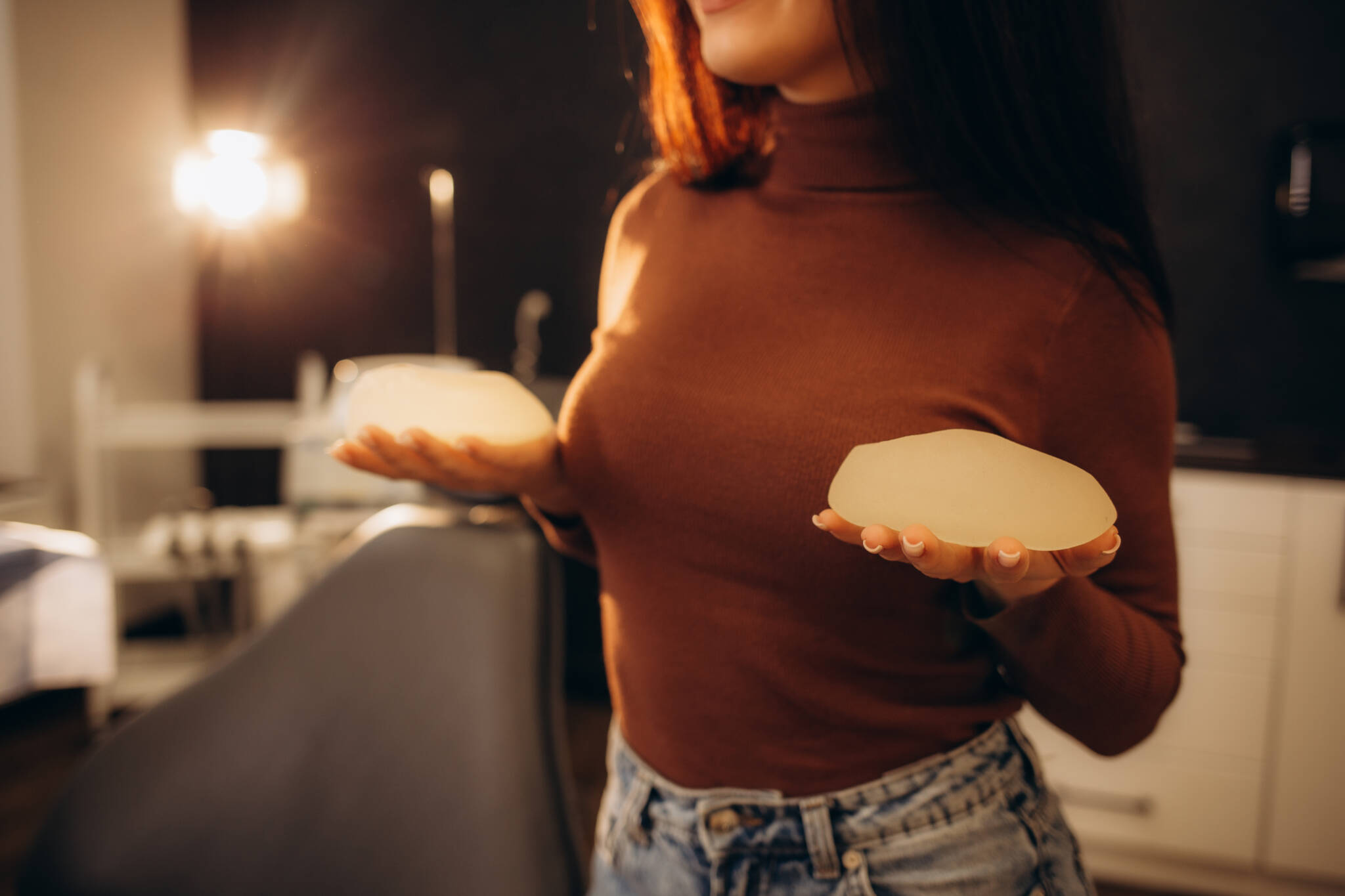 Woman wearing brown turtle neck holding silicone breast implants in each hand